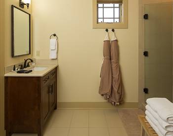 to the left is a dark cabinet with a light marble top with sink, bath robes handing on hooks with the glass inclosed shower with a ll tile. 